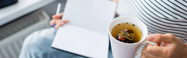 Colpo panoramico di donna che tiene una tazza di tisana e taccuino a casa — Foto stock