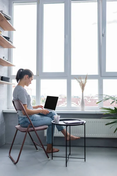 Seitenansicht eines barfüßigen Telearbeiters mit Laptop in der Nähe von Tasse und Notizbuch auf Couchtisch und Fenster zu Hause — Stockfoto