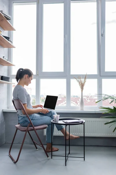 Side view of barefoot teleworker using laptop near cup and notebook on coffee table and window at home — Stock Photo