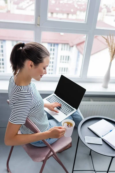 Morena freelancer utilizando el ordenador portátil mientras sostiene la taza de té de hierbas cerca de portátil y teléfono inteligente en la mesa - foto de stock