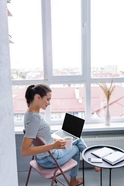 Selective focus of barefoot freelancer holding cup of herbal tea and laptop near smartphone and notebook on coffee table — Stock Photo