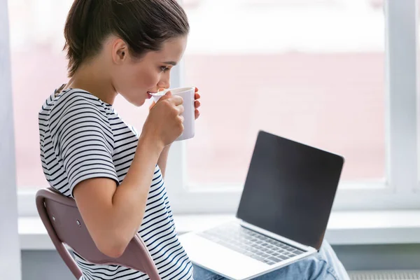 Selective focus of woman drinking tea and looking at laptop with blank screen at home — Stock Photo