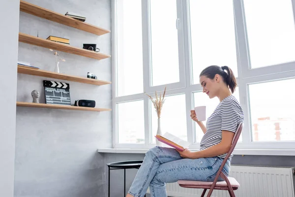 Vista laterale della donna con le gambe incrociate che tengono la tazza e leggono a casa — Foto stock