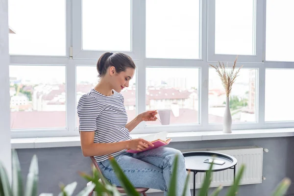 Selective focus of woman with cup reading book near smartphone and coffee table at home — Stock Photo