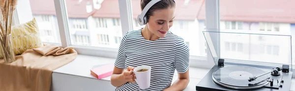 Panoramic shot of young woman in headphones holding cup near vinyl player on windowsill — Stock Photo