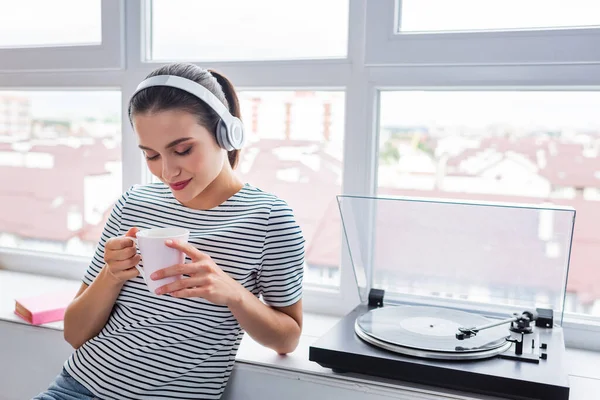 Junge Frau mit Kopfhörer hält Tasse neben Plattenspieler auf Fensterbank — Stockfoto