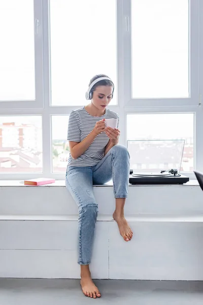 Donna a piedi nudi in cuffie in possesso di tazza mentre seduto vicino libro e vinile giocatore sul davanzale della finestra — Foto stock