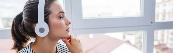 Foto panorâmica de mulher morena ouvindo música em fones de ouvido perto da janela — Fotografia de Stock