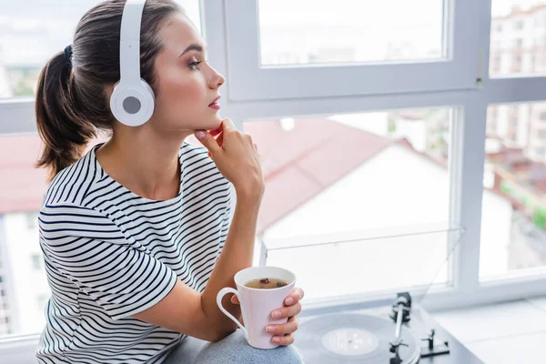 Mujer joven con taza de té escuchando música en auriculares cerca de reproductor de vinilo en el alféizar de la ventana - foto de stock