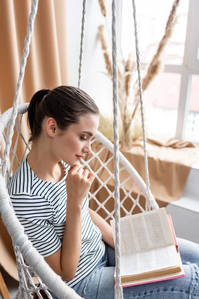Focus sélectif de la jeune femme livre de lecture dans le fauteuil suspendu à la maison — Photo de stock