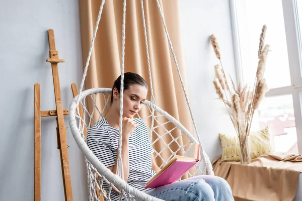 Young woman reading book while sitting in hanging armchair at home — Stock Photo