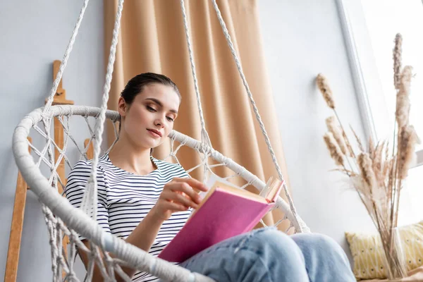 Selektiver Fokus der brünetten Frau, die Buch im hängenden Sessel im Wohnzimmer liest — Stockfoto