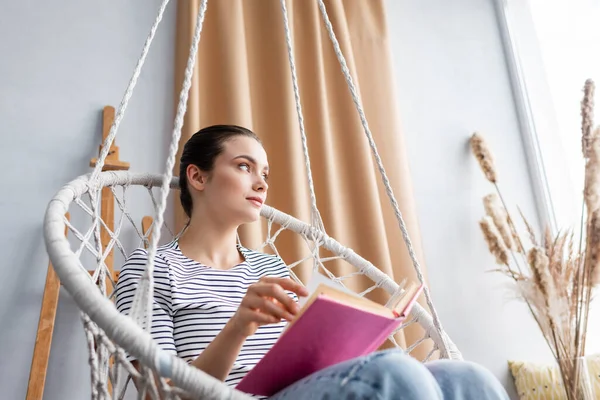 Enfoque selectivo de la mujer joven sosteniendo libro mientras está sentada en un sillón colgante - foto de stock