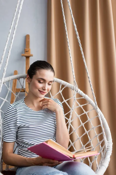 Selective focus of brunette woman reading book in hanging armchair at home — Stock Photo