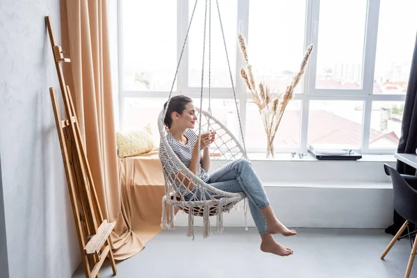 Mujer joven con los ojos cerrados sosteniendo la taza mientras está sentado en el sillón colgante - foto de stock