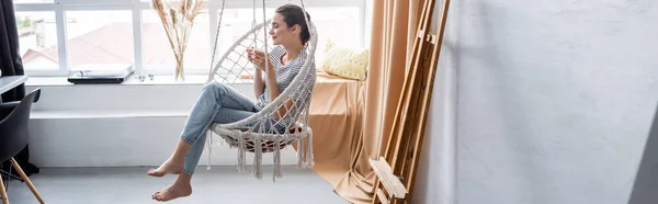 Orientação panorâmica da mulher descalça segurando copo enquanto sentada na poltrona pendurada em casa — Fotografia de Stock