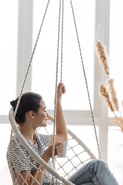 Selective focus of brunette woman holding cup while sitting in hanging armchair at home — Stock Photo