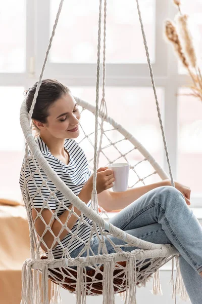 Selektiver Fokus einer brünetten Frau, die zu Hause eine Tasse im hängenden Sessel hält — Stockfoto