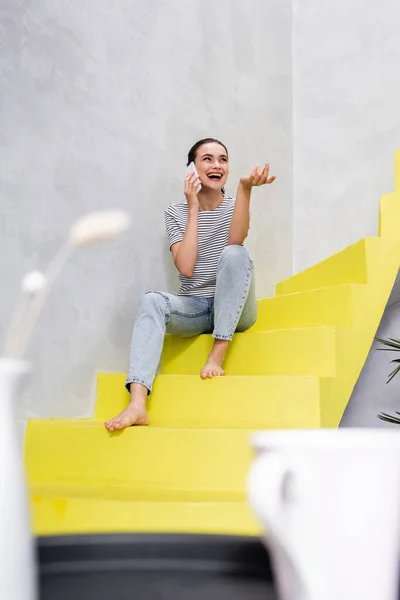 Focus selettivo della donna eccitata che parla sullo smartphone mentre è seduta sulla scala a casa — Foto stock