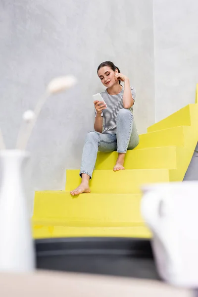 Селективный фокус босиком женщины с помощью смартфона, сидя на лестнице дома — стоковое фото