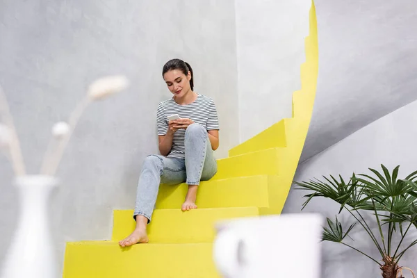 Selective focus of barefoot woman using smartphone on stairs at home — Stock Photo