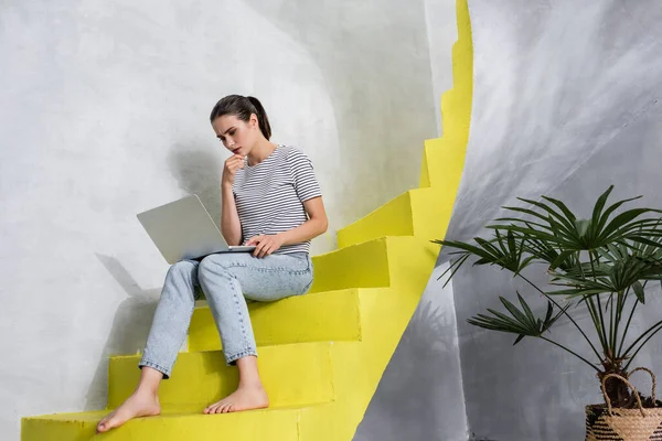 Femme pensive travaillant sur un ordinateur portable tout en étant assis sur des escaliers à la maison — Photo de stock