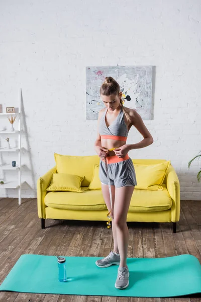 Mujer deportiva delgada midiendo la cintura con cinta de pie en la esterilla de fitness en casa - foto de stock