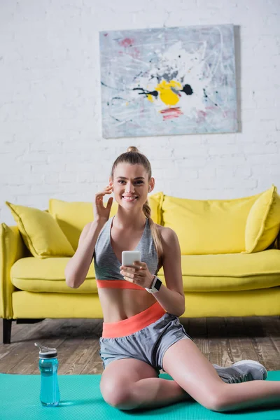 Vue de face de la sportive assise sur un tapis de fitness près d'une bouteille de sport à la maison — Photo de stock