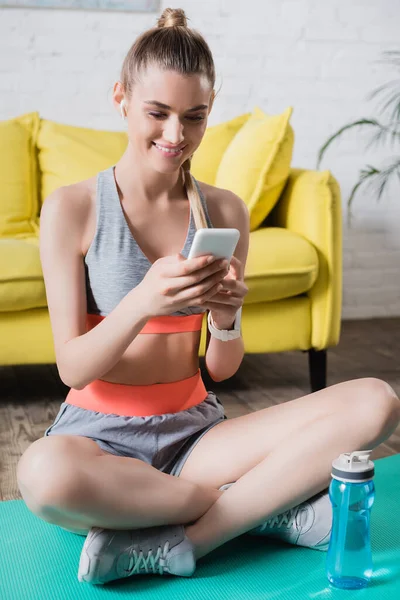 Deportiva con teléfono celular sentada cerca de la botella deportiva en la colchoneta de fitness en casa - foto de stock