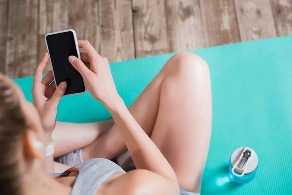 Enfoque selectivo de la deportista con teléfono celular sentado en la alfombra de fitness en casa - foto de stock