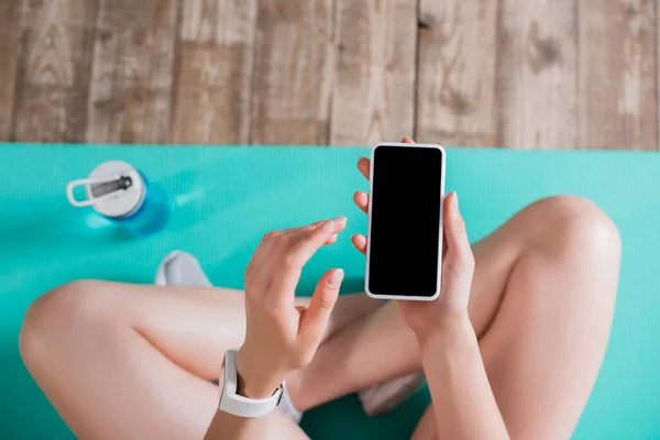 Vista superior de la mujer con teléfono celular con pantalla en blanco cerca de la botella de deportes en casa - foto de stock