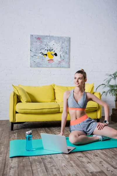 Sportswoman sitting on fitness mat near laptop and sports bottle at home — Stock Photo