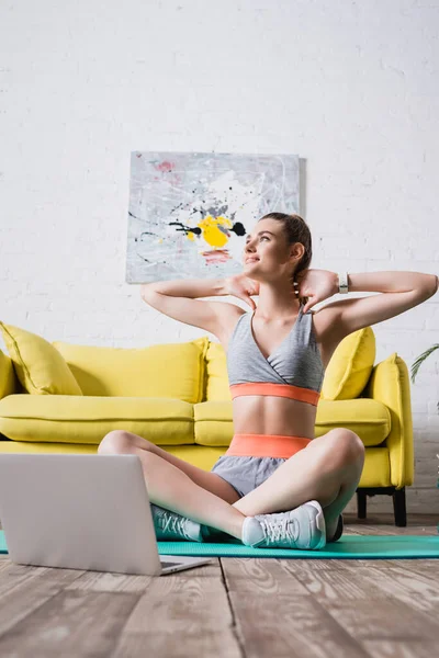 Desportista com as mãos atrás da cabeça sentado perto do laptop no tapete de fitness em casa — Fotografia de Stock