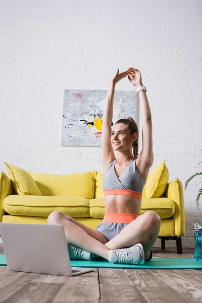 Sportiva con gambe incrociate che si estende vicino al laptop sul tappeto fitness a casa — Foto stock