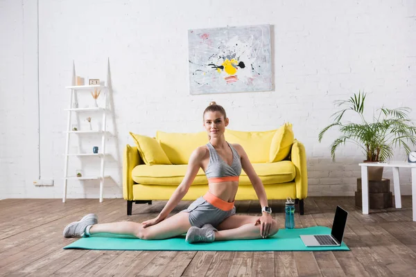 Sportswoman formation près de l'ordinateur portable et bouteille de sport sur tapis de fitness à la maison — Photo de stock