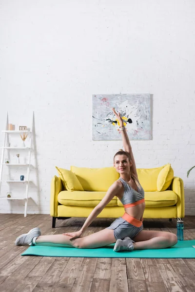 Deportiva con la mano en el aire haciendo ejercicio cerca de la botella de deportes en la alfombra de fitness en casa - foto de stock