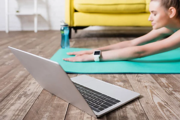 Concentration sélective d'un ordinateur portable près d'une sportive s'étirant sur un tapis de fitness à la maison — Photo de stock