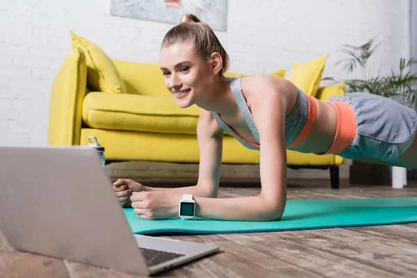 Selective focus of smiling sportswoman standing in plank near laptop at home — Stock Photo