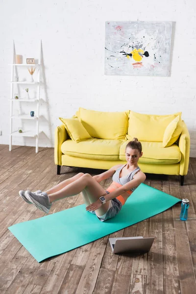 Giovane sportiva che si esercita sul tappeto fitness vicino al laptop e alla bottiglia sportiva a casa — Foto stock