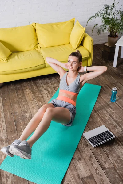 Vue en angle élevé de la sportive gaie travaillant sur tapis de fitness près d'un ordinateur portable et bouteille de sport à la maison — Photo de stock