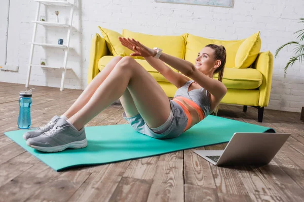 Selective focus of smiling sportswoman training on fitness mat near sports bottle and laptop — Stock Photo