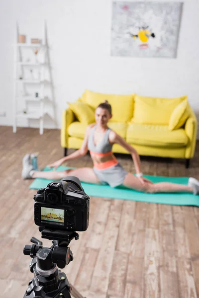 Focus selettivo della giovane sportiva che fa split sul tappeto fitness vicino alla fotocamera digitale a casa — Foto stock