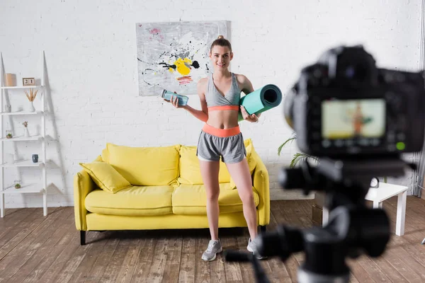 Selective focus of smiling sportswoman holding sports bottle and fitness mat near digital camera — Stock Photo