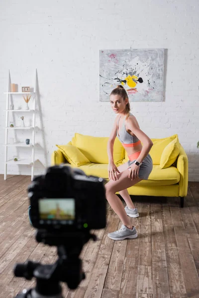 Selective focus of young woman in sportswear doing squat near digital camera in living room — Stock Photo