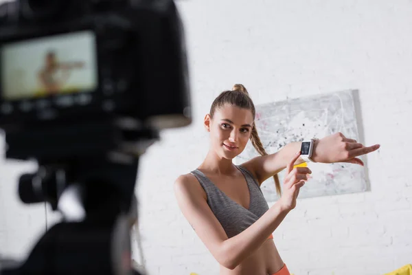 Selective focus of smiling sportswoman pointing at smart watch near digital camera at home — Stock Photo