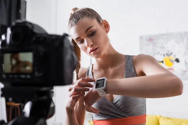 Enfoque selectivo de la mujer en ropa deportiva apuntando al rastreador de fitness cerca de la cámara digital en casa - foto de stock