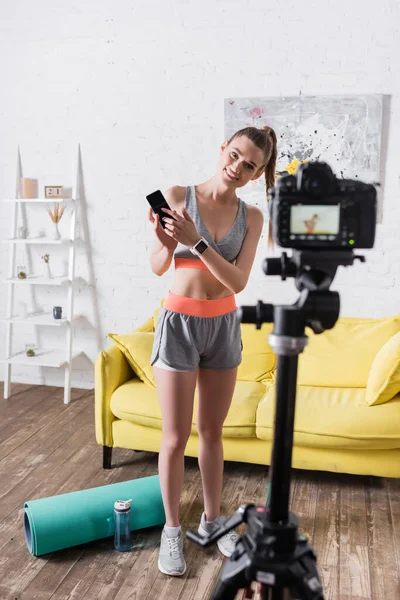 Selective focus of smiling sportswoman pointing at smartphone near digital camera and fitness mat at home — Stock Photo