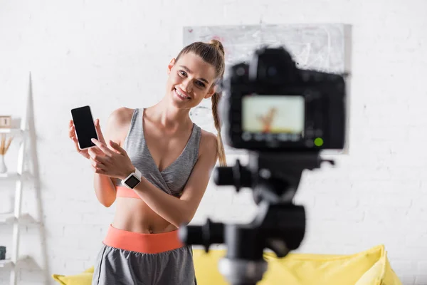 Selective focus of positive sportswoman pointing at smartphone near digital camera at home — Stock Photo