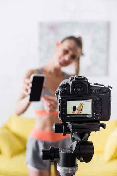 Focus selettivo della donna sorridente in abbigliamento sportivo con smartphone con schermo bianco vicino alla fotocamera digitale a casa — Foto stock