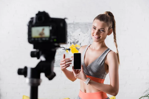 Enfoque selectivo de la deportista positiva mirando a la cámara digital mientras sostiene el teléfono inteligente con pantalla en blanco - foto de stock